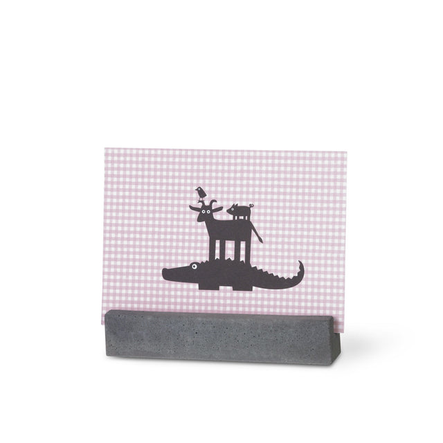 Ein Bilderhalter aus Beton mit einer Postkarte, die ein humorvolles Motiv zeigt: Ein Krokodil, eine Ziege und ein Schwein sind dargestellt.
