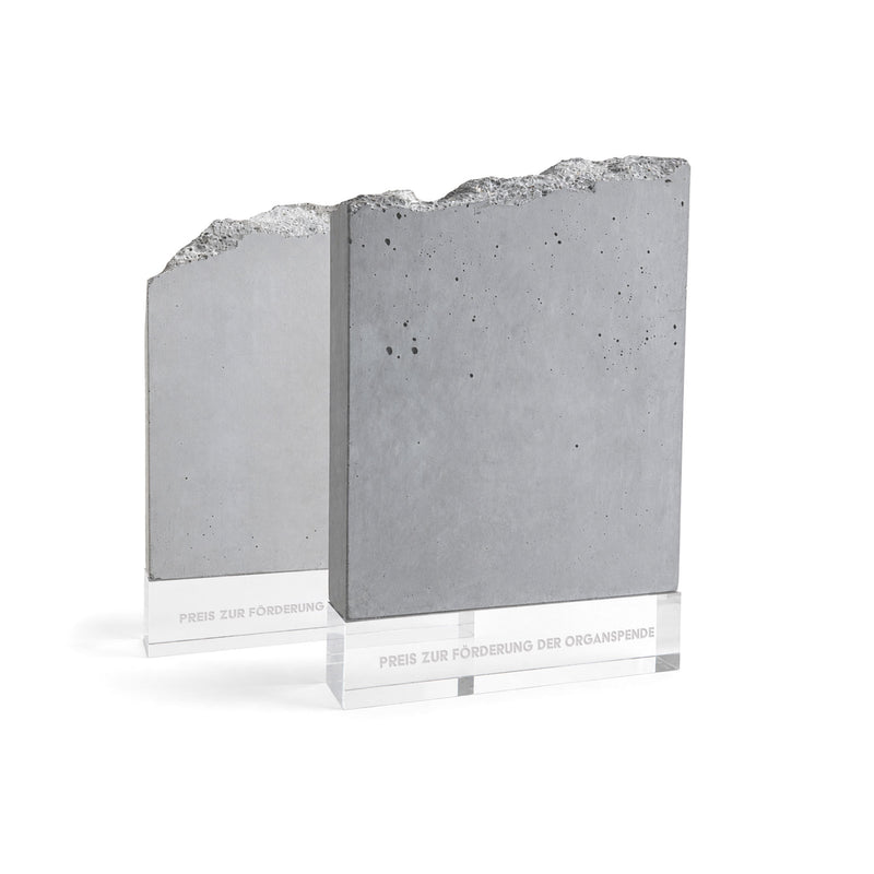 Zwei Beton-Awards mit roh gebrochener Kante auf transparentem Glasfuß, gravierter Schriftzug