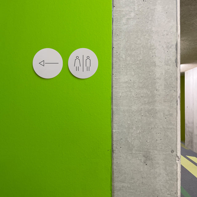 Zwei runde Betonschilder mit den Piktogrammen Toilette und Pfeil nach links
